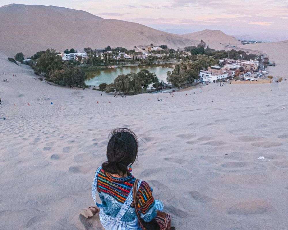 A person sits atop a sand dune, overlooking the lush oasis of Huacachina, surrounded by golden desert sands, one of the remarkable places to visit in Peru.