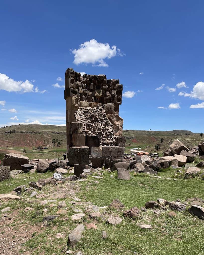 A partially ruined stone tower stands under a clear blue sky at the archaeological site of Sillustani, surrounded by scattered rocks and green fields, highlighting one of the fascinating places to visit in Peru.