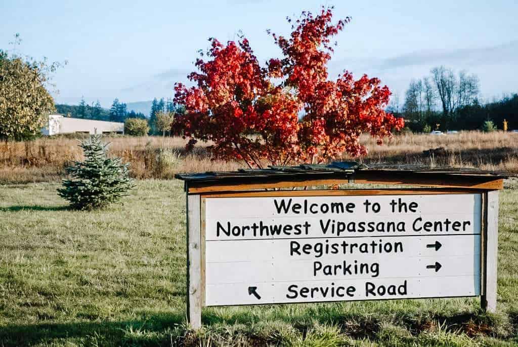 A sign welcomes visitors to the Northwest Vipassana Center, guiding them to registration, parking, and the service road. The sign is set against a backdrop of open fields and a vibrant red tree, emphasizing the peaceful setting ideal for a Vipassana Meditation Retreat.