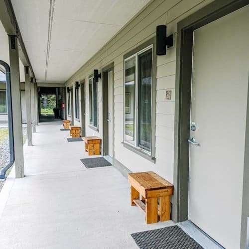 The exterior of the accommodation area at a Vipassana Meditation Retreat, featuring a long, covered walkway with several doors leading to individual rooms. Small wooden benches are placed in front of each door. The clean, minimalist design emphasizes a serene and quiet environment, ideal for meditative practice.