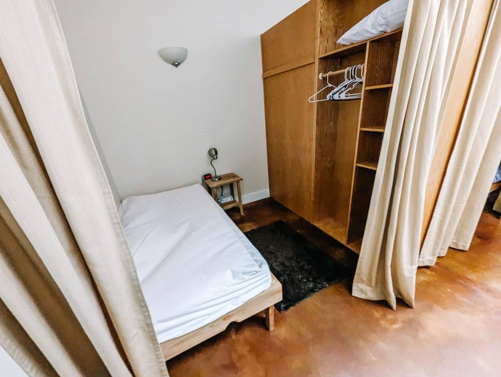 A simple and tidy accommodation at a Vipassana Meditation Retreat, featuring a single bed with white linens, a small bedside table with a lamp, and an open wardrobe with shelves and hangers, all separated by light-colored curtains.