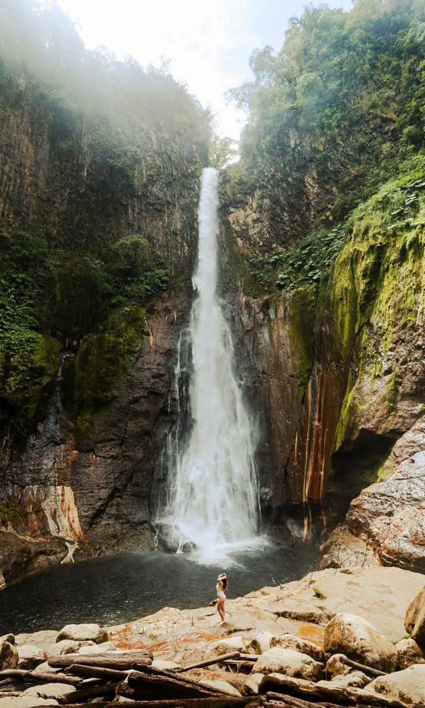 A person stands in awe at the base of a towering waterfall surrounded by lush, green cliffs during one of the Costa Rica day trips, capturing the dramatic beauty of the natural landscape.