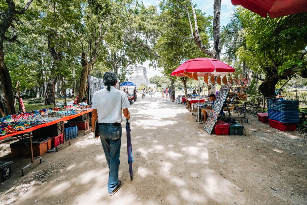 A visitor walking through a lively path lined with souvenir stalls under red umbrellas during Chichen Itza day trips, with trees providing shade along the way.