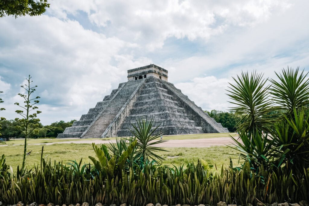 The El Castillo pyramid at Chichen Itza day trips stands tall on a sunny day, framed by green plants in the foreground and a partly cloudy sky above.