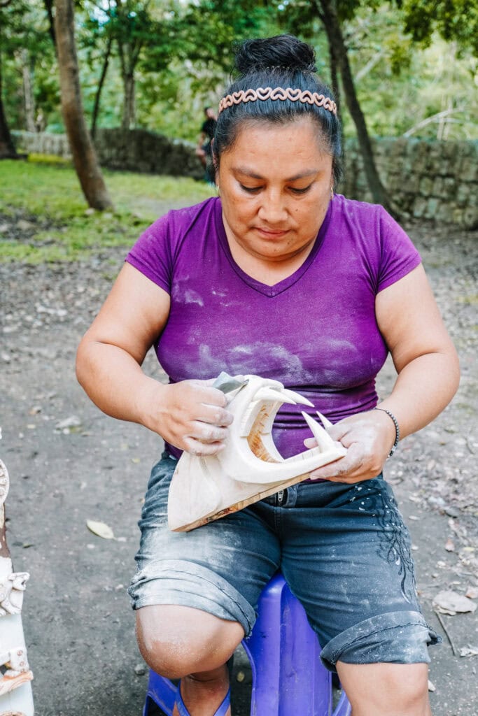 Local artisan shaping a small craft during Chichen Itza day trips, sitting on a blue stool and focused on her work.