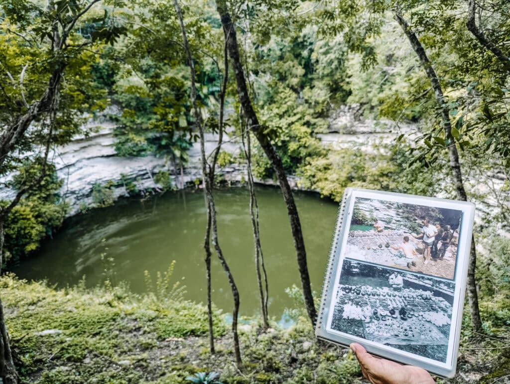 I'm holding a photo book overlooking the Sacred Cenote during Chichen Itza day trips, with the green water surrounded by thick trees.